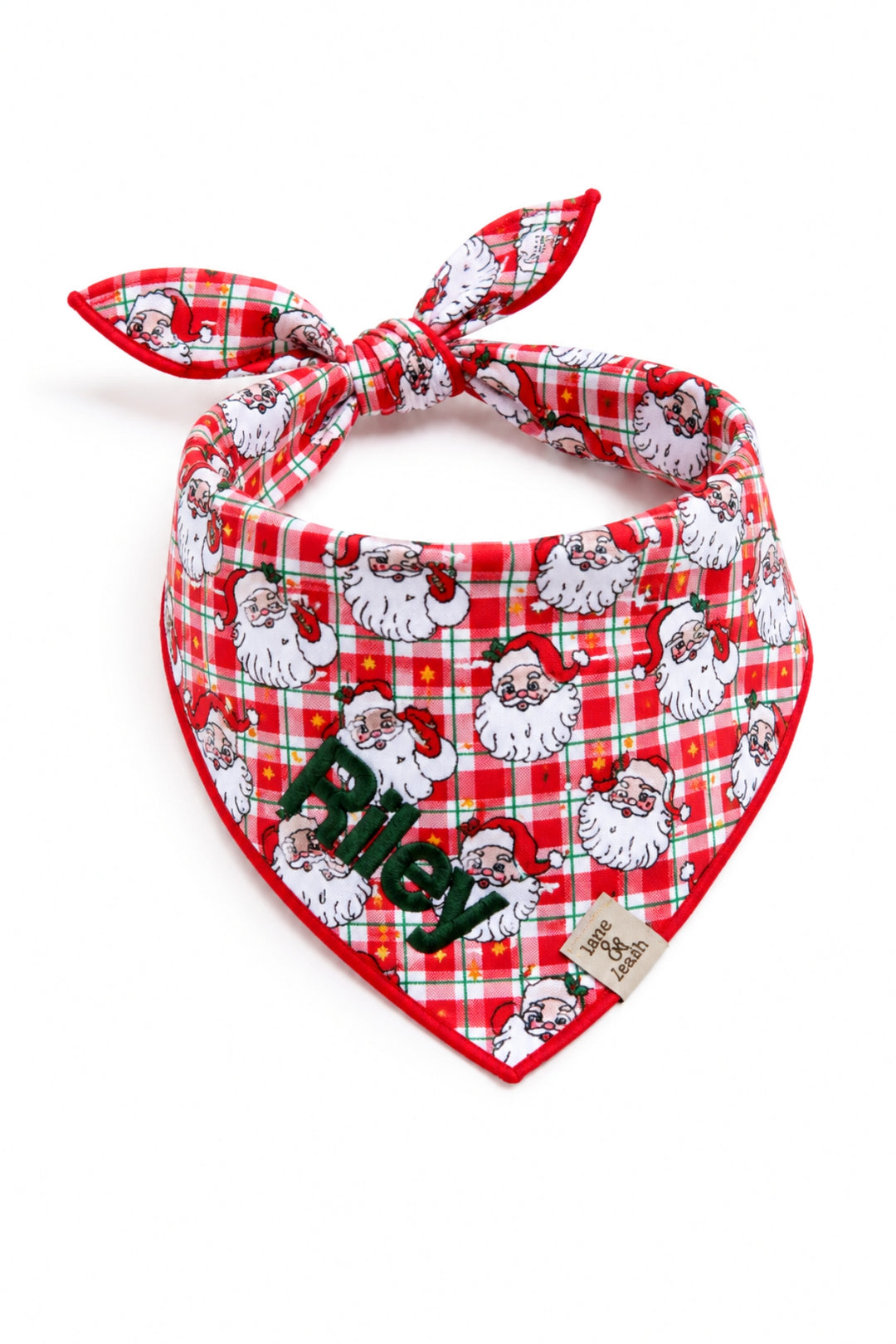A red and white plaid dog bandana from Lane & Leash tied in a knot, featuring a repeating pattern of vintage-style Santa Claus faces with white beards and red hats, accented with thin green grid lines and small gold stars. The name 'Riley' is embroidered in dark green block lettering near the bottom point, with a small woven Lane & Leash label tag