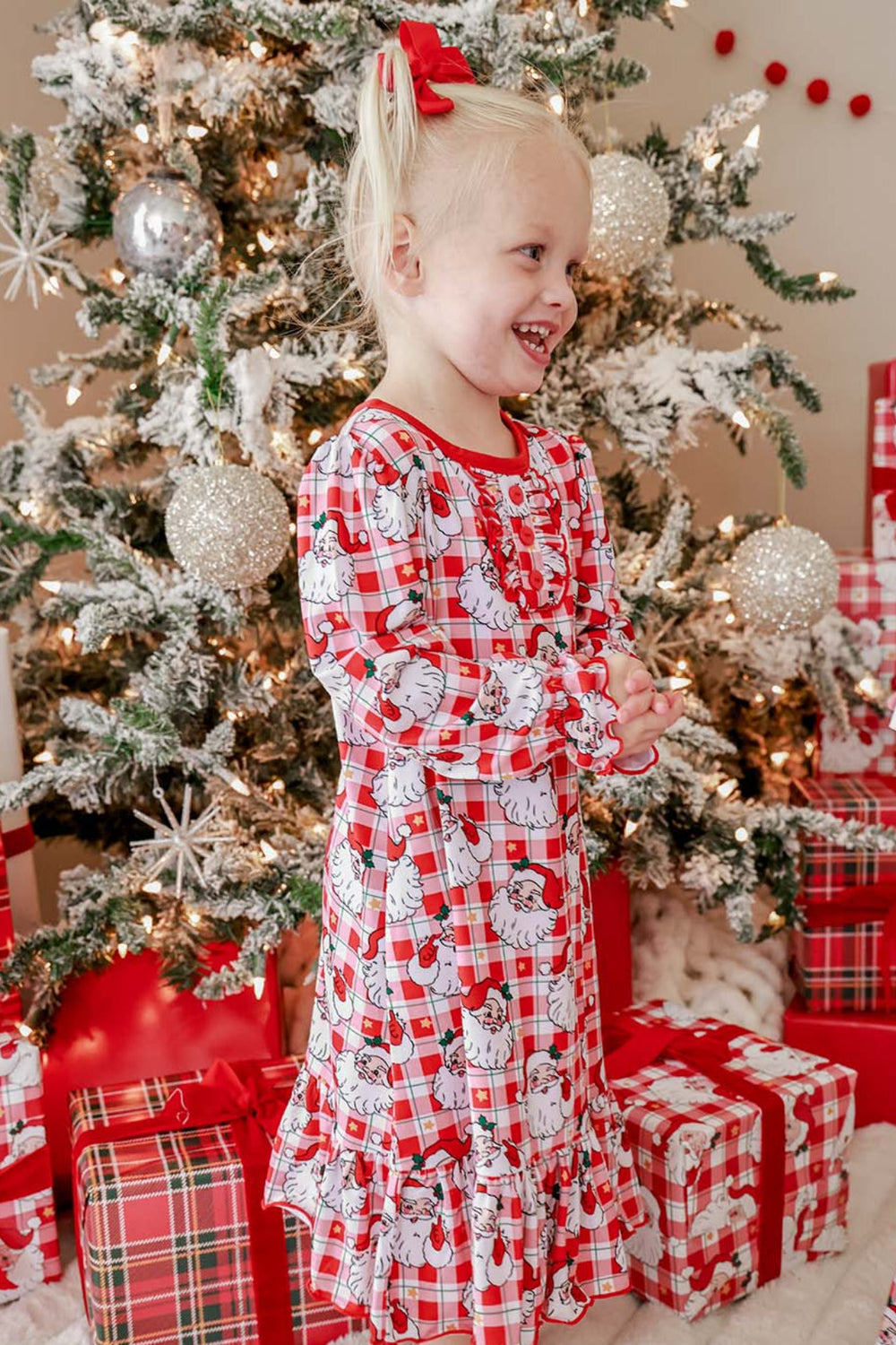 Little girl wearing a Caden Lane Cozy Claus Christmas princess ruffle nightgown in a vintage Santa plaid print beside a decorated Christmas tree.