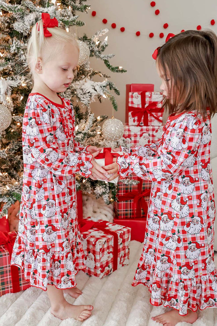 Matching friends wearing Caden Lane Cozy Claus Christmas princess ruffle nightgowns in a vintage Santa plaid print while exchanging a holiday gift by the Christmas tree.