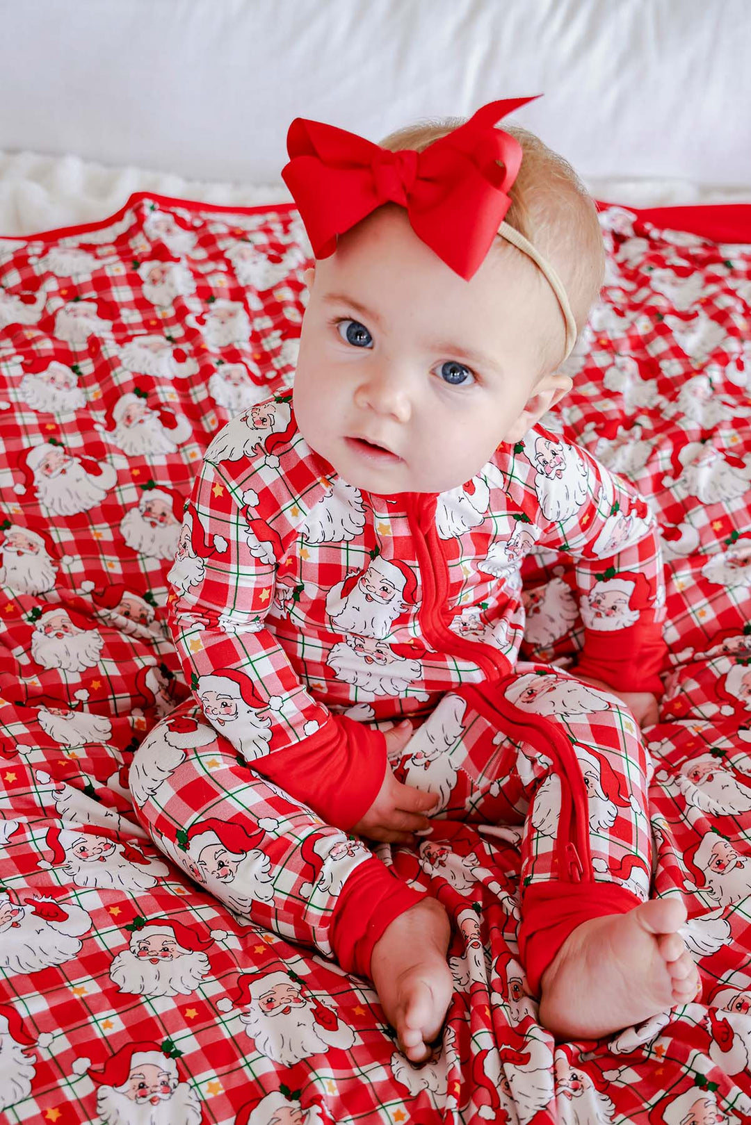 Baby wearing a Caden Lane Cozy Claus Christmas bamboo zipper romper featuring a vintage Santa plaid print, sitting on a matching holiday blanket with a festive red bow headband.