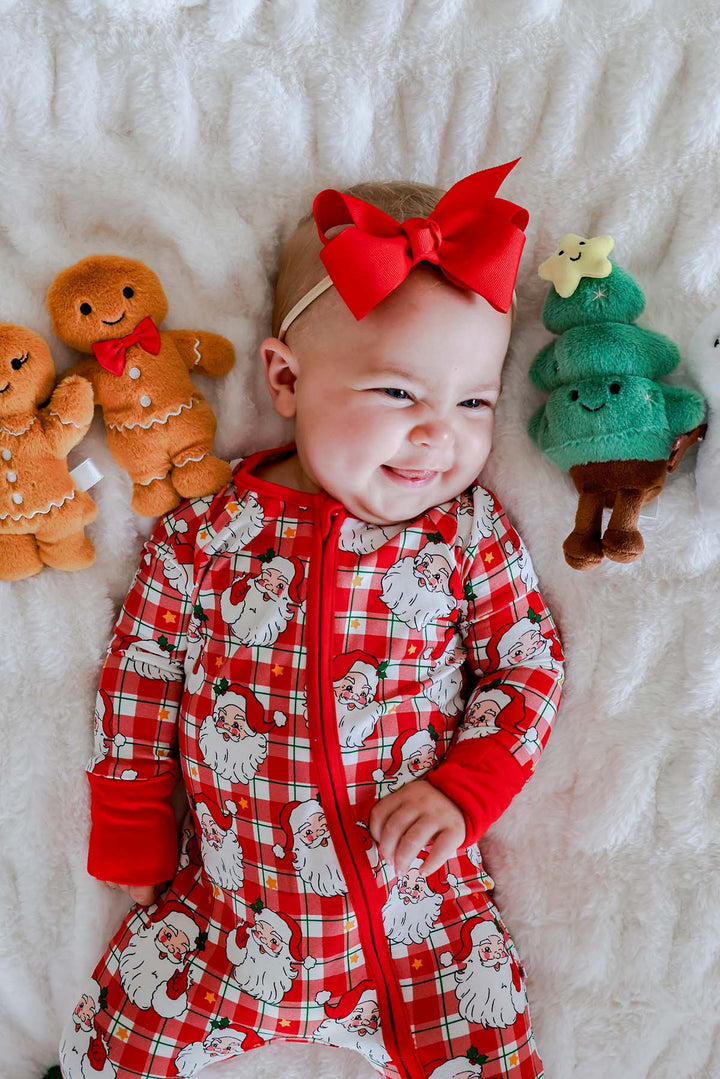 A baby lying on a white textured blanket, wearing a red and white Santa plaid zip-up onesie and a red bow headband. Gingerbread and Christmas tree plush toys are placed on either side of the baby.