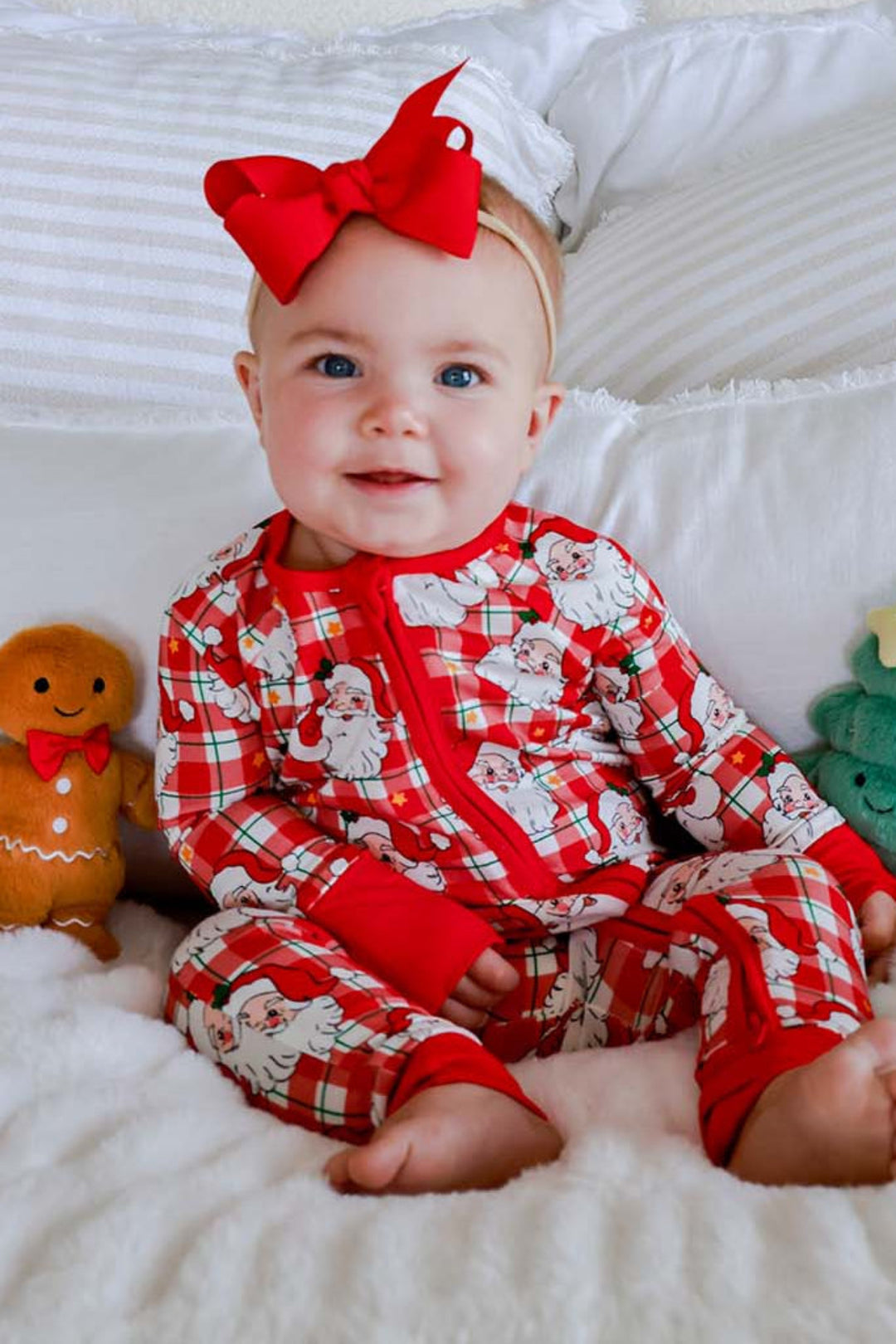 A baby sitting on a white blanket in front of white pillows, wearing red and white holiday pajamas with a large red bow in their hair. The scene is decorated with gingerbread cookies, a snowman toy, and a green wreath on the wall.
