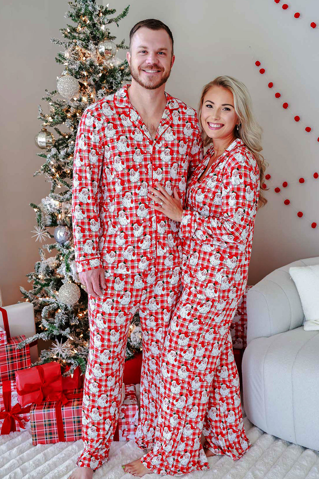 A smiling couple wearing matching red and white Santa Claus patterned pajamas, standing barefoot in front of a decorated Christmas tree with wrapped gifts and a light-colored sofa.