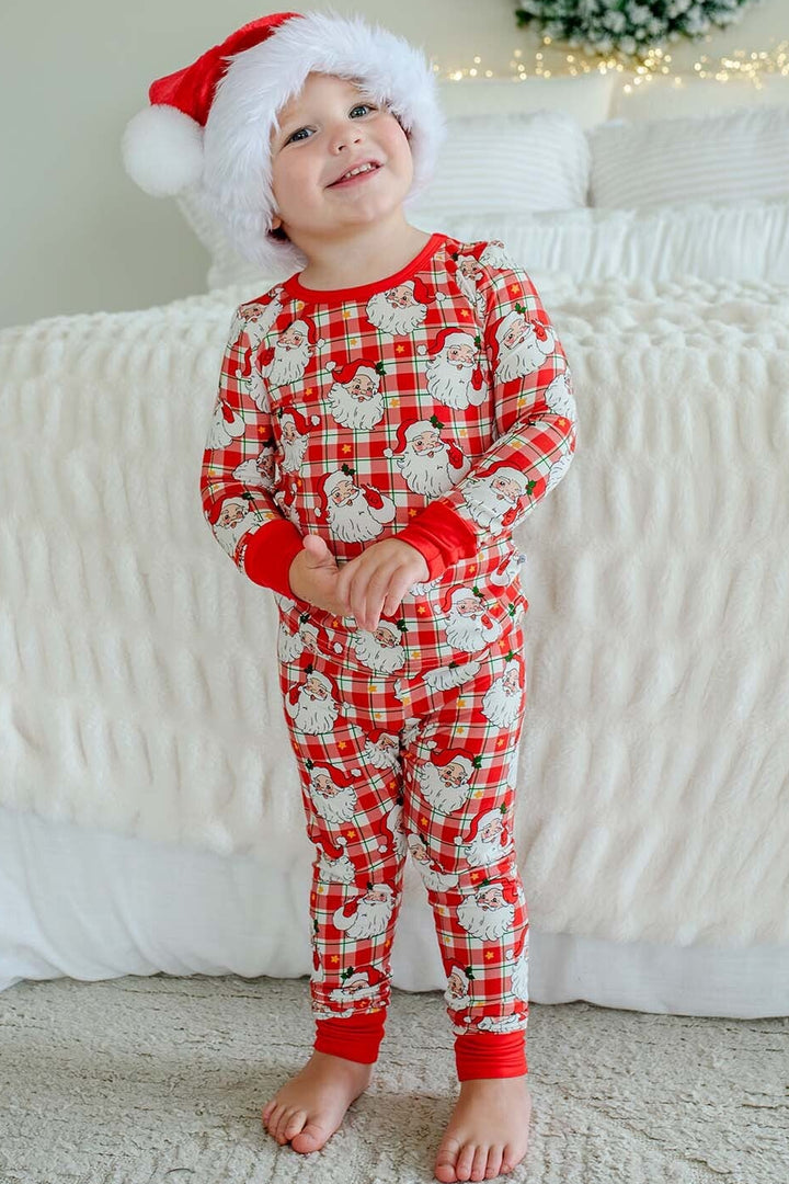 A toddler wearing a red and white Santa-themed pajama set with a matching Santa hat, standing barefoot in front of a white bed with a holiday wreath in the background.
