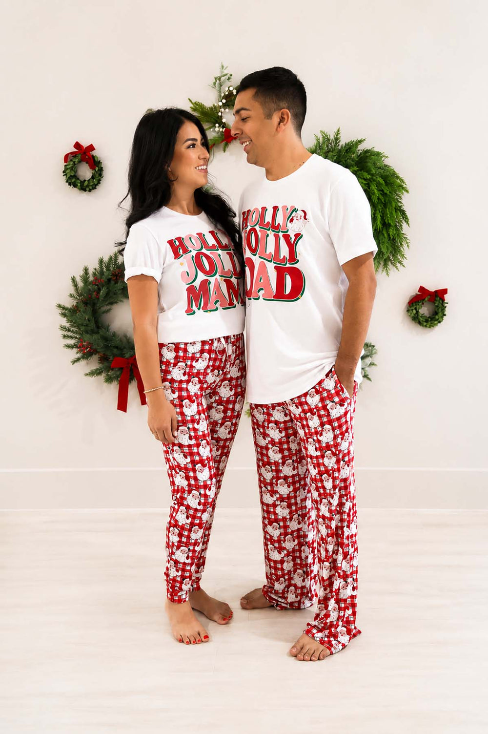 Couple wearing matching Caden Lane "Holly Jolly Mama" and "Holly Jolly Dad" white t-shirts with red plaid Santa-print pajama pants, standing together in front of a wreath-decorated wall.