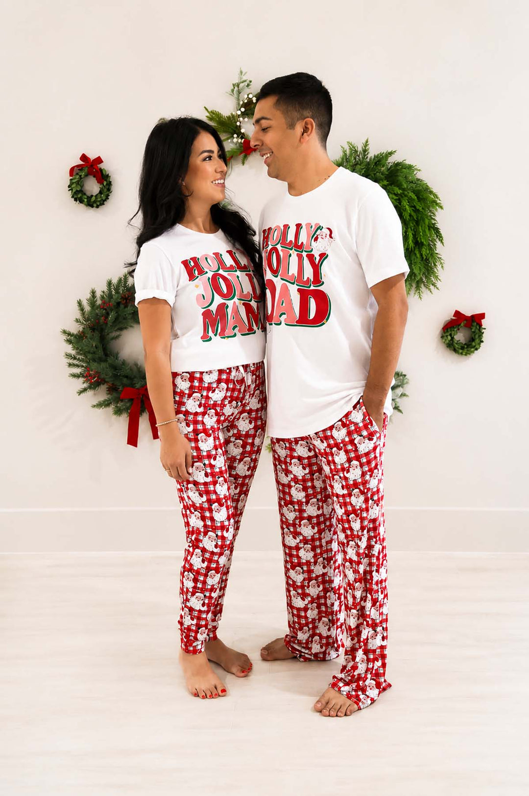 Couple wearing matching Caden Lane "Holly Jolly Mama" and "Holly Jolly Dad" white t-shirts with red plaid Santa-print pajama pants, standing together in front of a wreath-decorated wall.