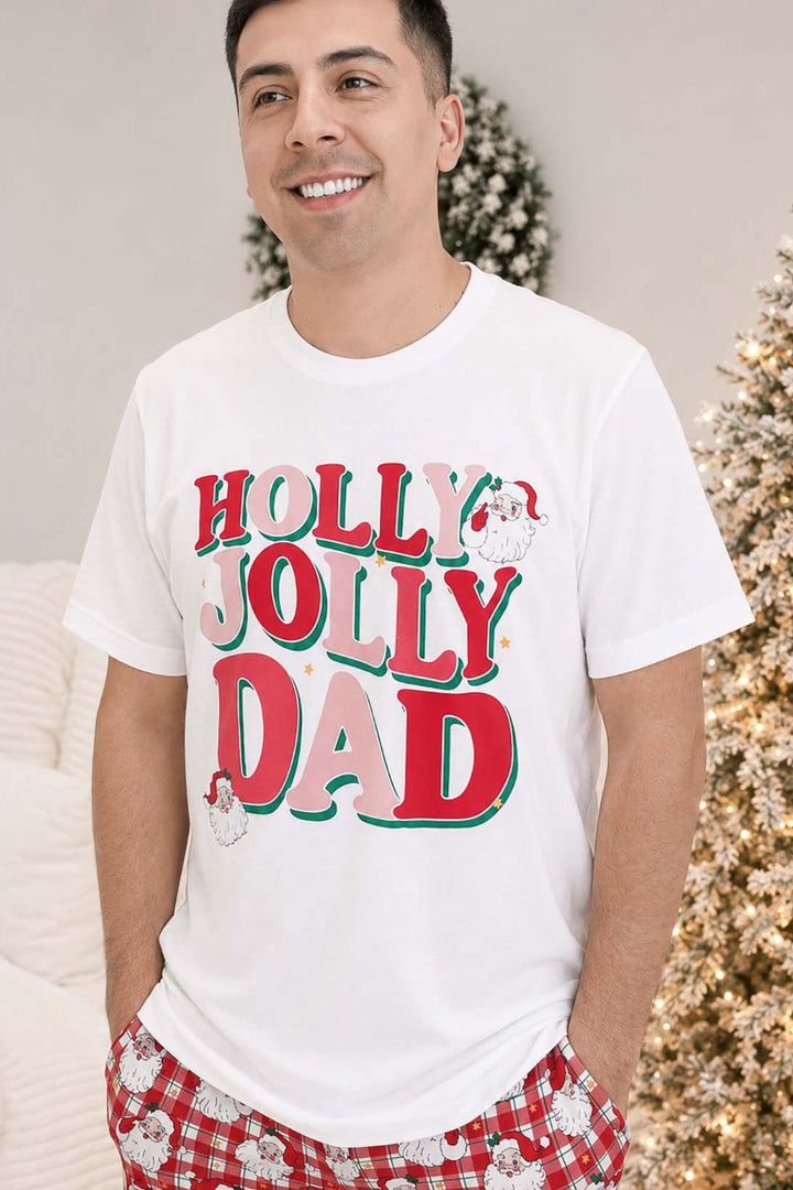 Man wearing a white Caden Lane "Holly Jolly Dad" graphic t-shirt with Santa illustrations, paired with red plaid Santa-print pajama pants, standing near a Christmas tree.