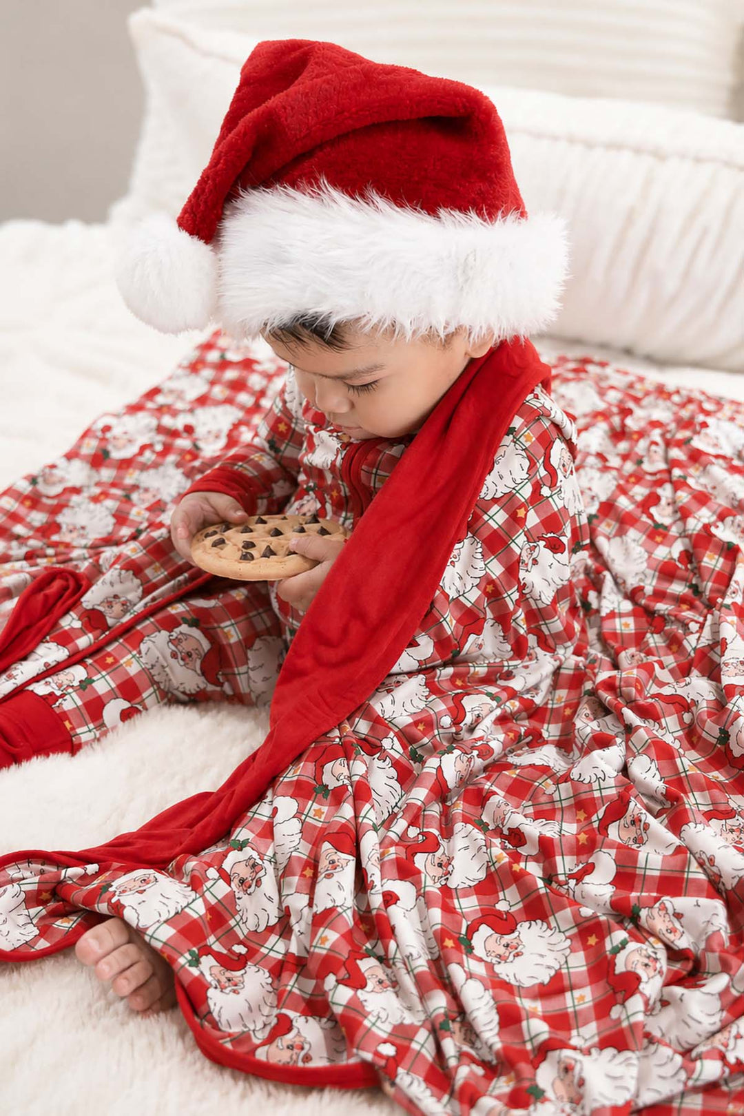 Toddler in Cozy Claus Christmas bamboo pajamas wearing a Santa hat and cuddling a personalized vintage Santa Christmas blanket while enjoying holiday cookies.