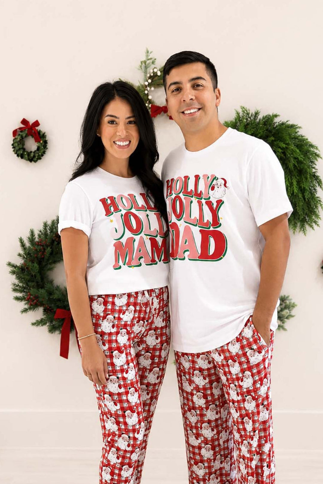 Couple smiling together in matching Caden Lane "Holly Jolly Mama" and "Holly Jolly Dad" t-shirts paired with red plaid Santa-print pajama pants, standing in front of Christmas wreaths.