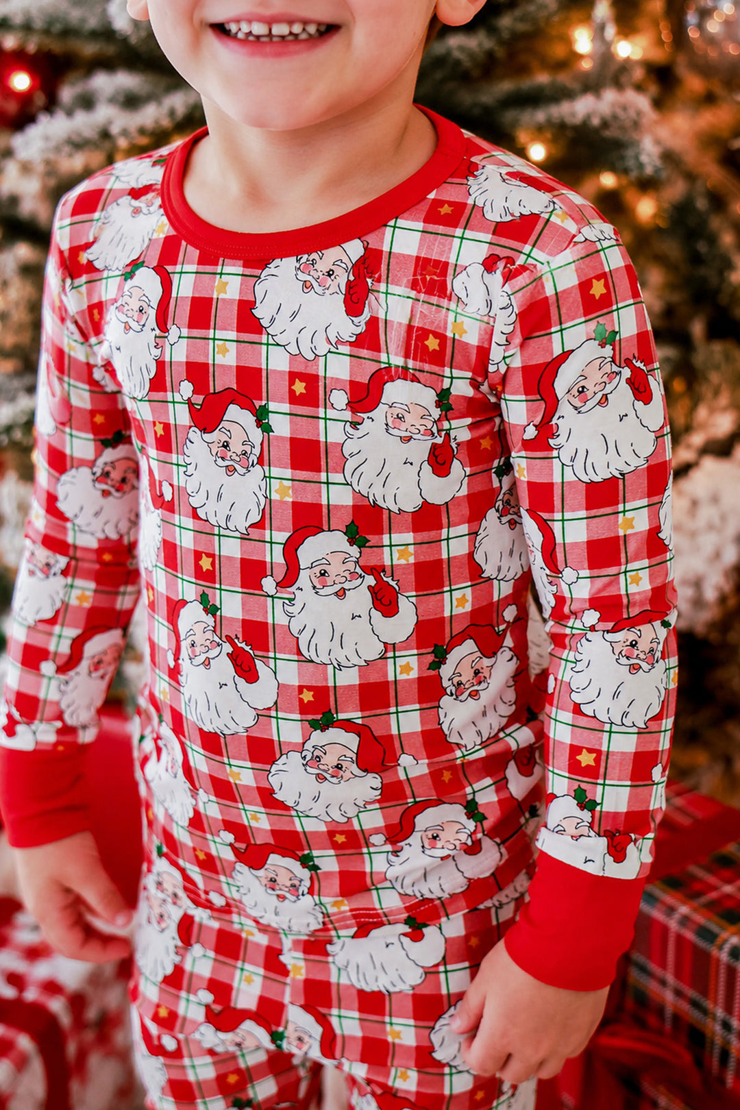 A child wearing red and white plaid pajamas with a repeating Santa Claus print, standing in front of a decorated Christmas tree.