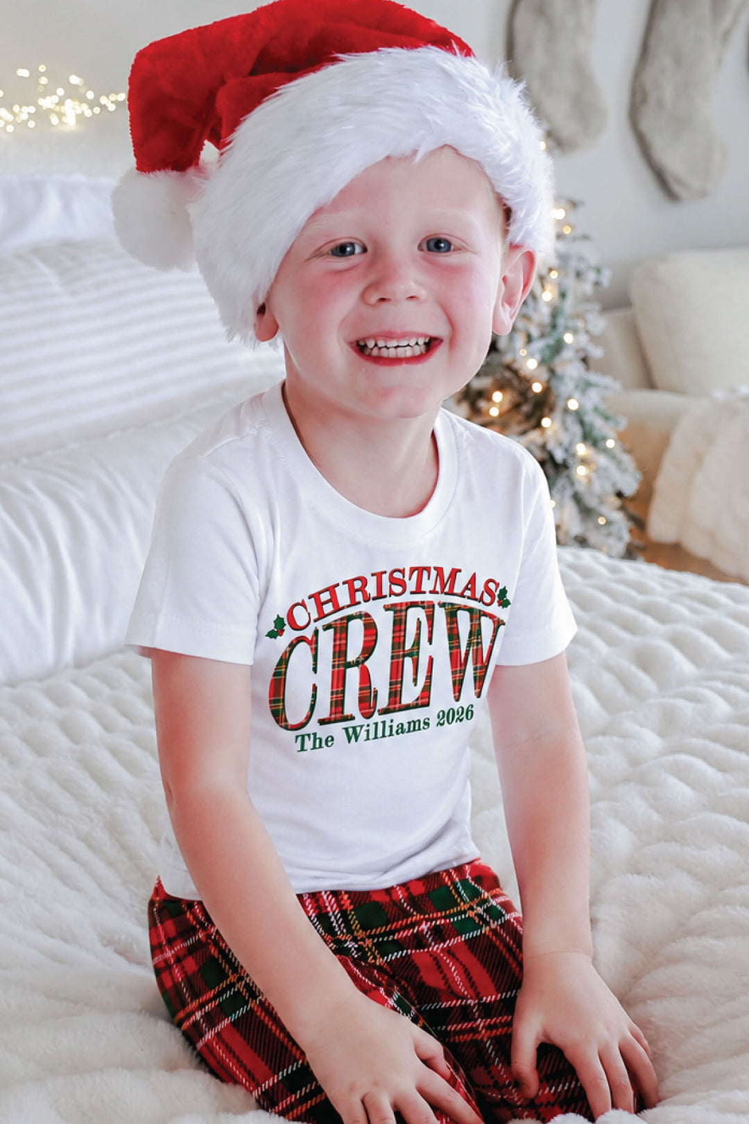 Smiling boy wearing a personalized Caden Lane Christmas Crew shirt with red plaid pajama pants and a Santa hat.