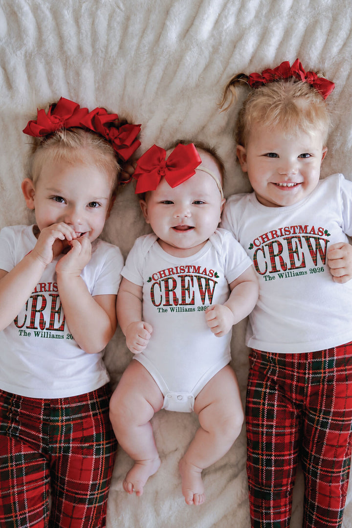 Three siblings wearing personalized Caden Lane Christmas Crew shirts and coordinating red plaid Christmas pajamas with festive red bows.