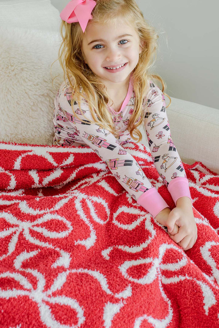 Caden Lane red bow print sherpa blanket laid over a young girl wearing pink nutcracker-print pajamas, showcasing the plush texture and allover ribbon bow pattern.