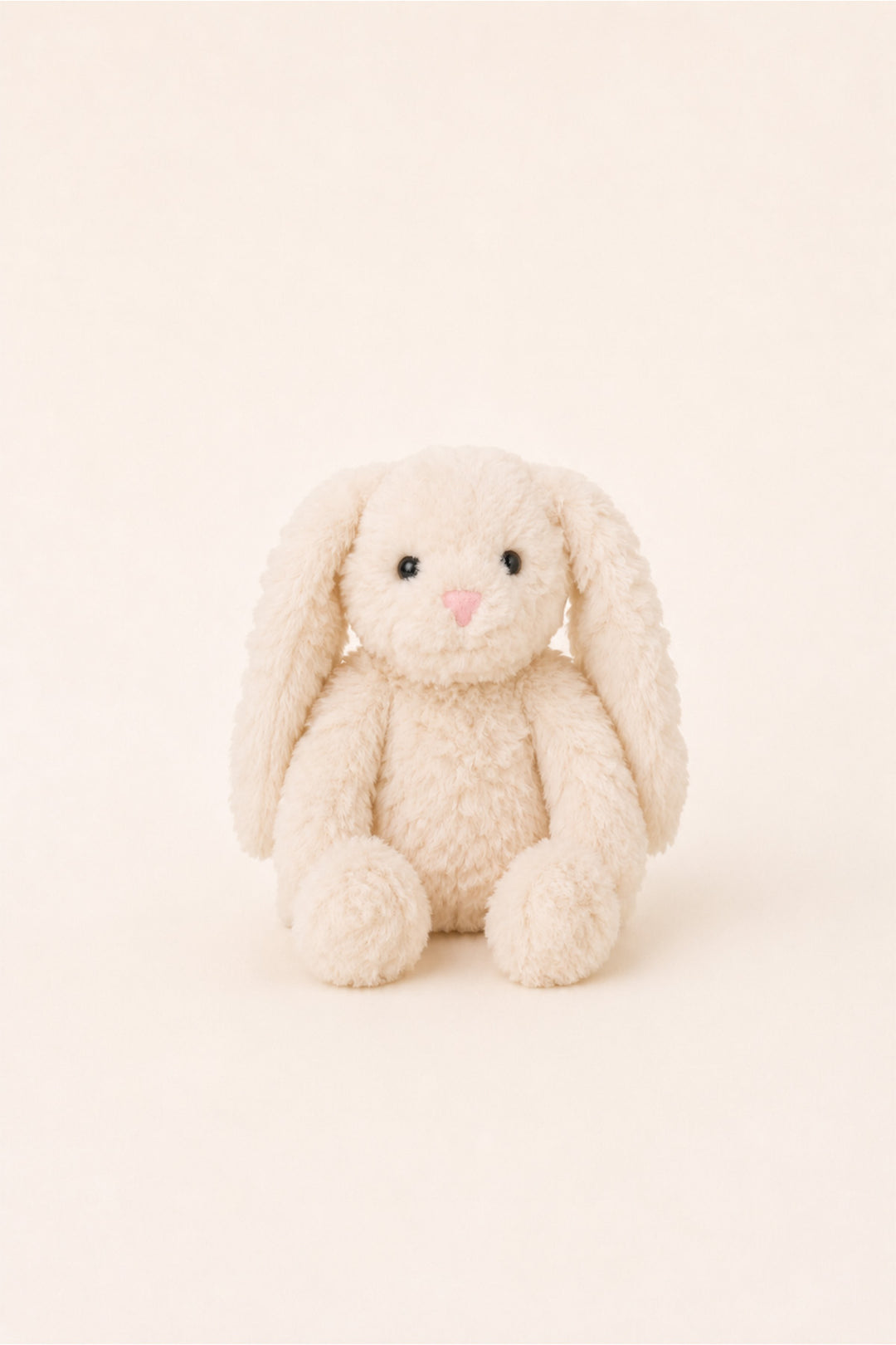 A cream-colored plush bunny toy with long floppy ears, black bead eyes, and a small pink nose, sitting against a solid cream background.