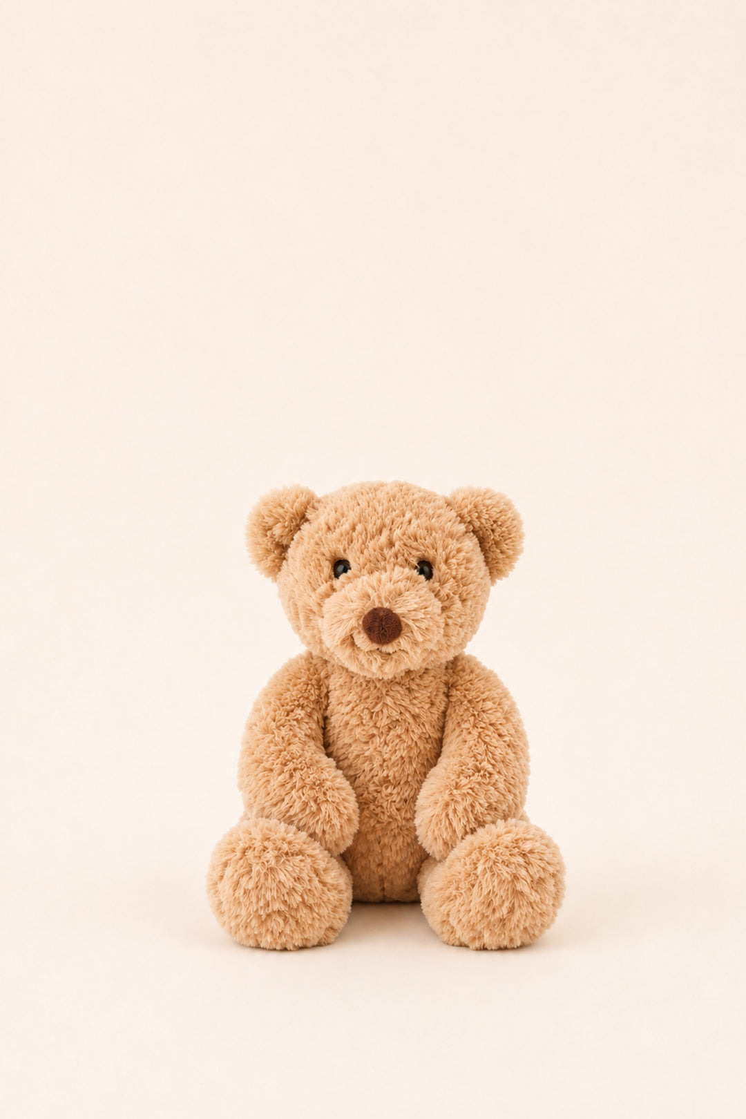 A small plush teddy bear in a warm tan color sits centered against a plain, light cream background.
