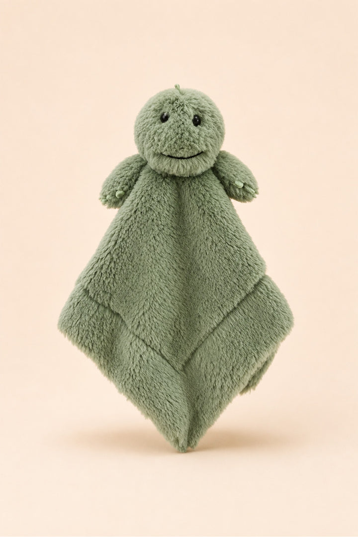 A Caden Lane plush dinosaur security blanket in a dusty sage green color, featuring a soft textured body, small embroidered eyes, and a stitched smile, set against a solid pale peach background.
