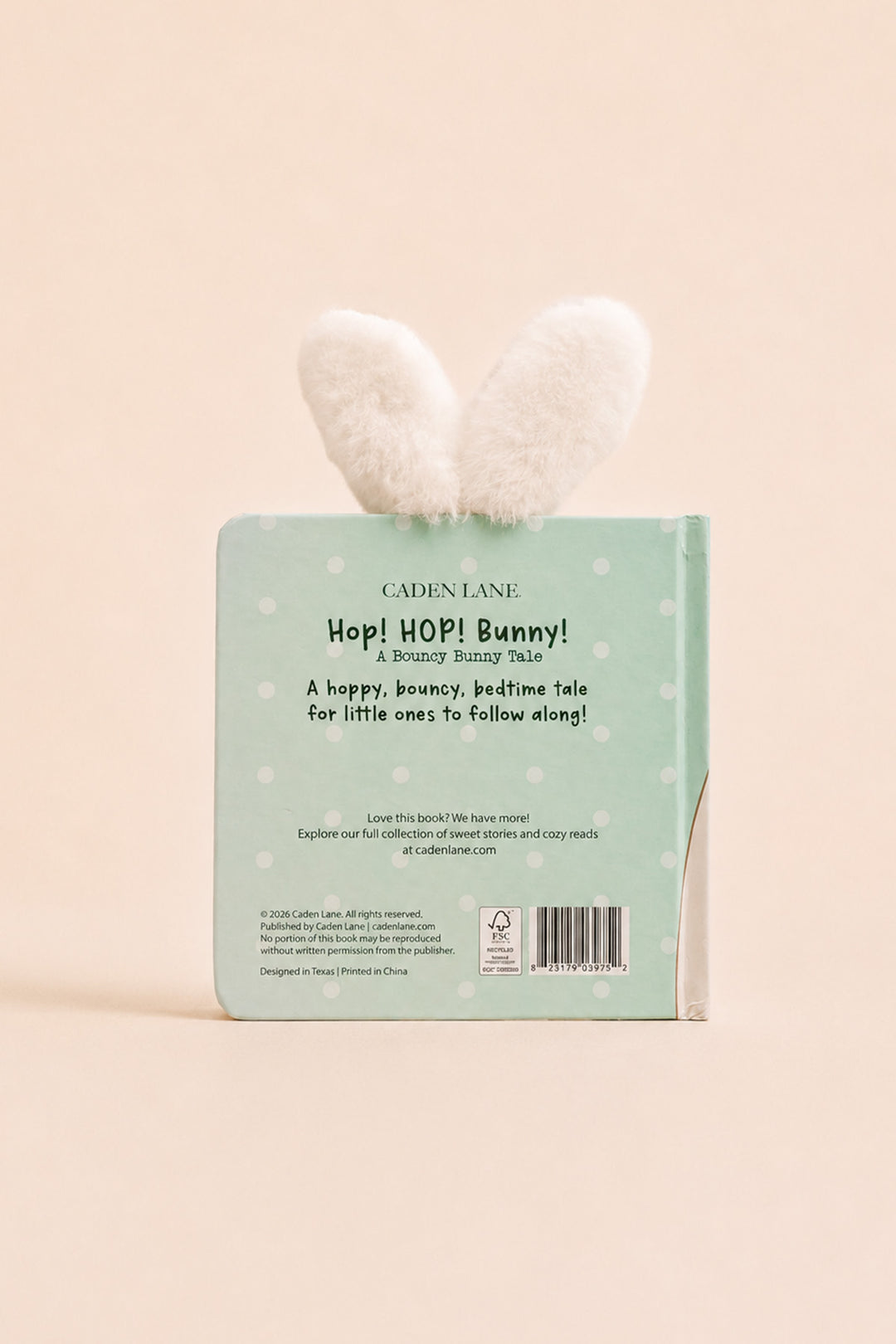 The back cover of the children's book 'Hop! Hop! Bunny!' by Cadence Lane, featuring a mint green background with white polka dots and a fluffy white bunny ear bookmark attached at the top, set against a solid pale peach background.
