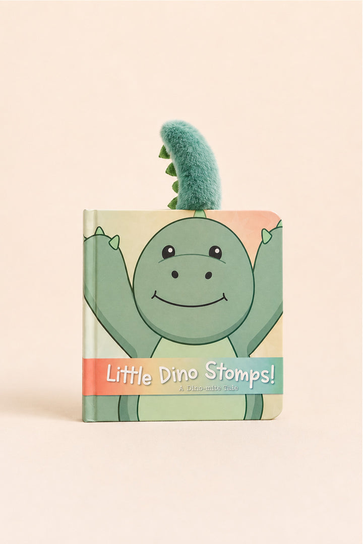 A children's book titled 'Little Dino Stomps!' featuring a smiling green cartoon dinosaur on the cover, with a small plush green dinosaur tail sticking out from the top, photographed on a soft beige background.