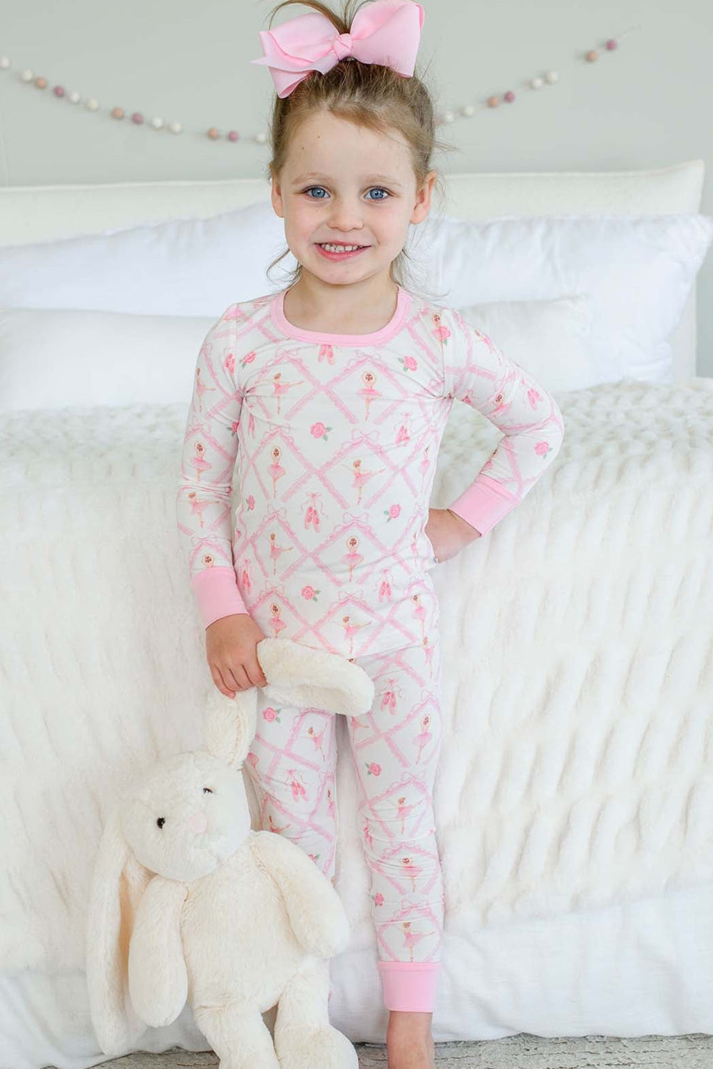 Smiling toddler girl wearing a Caden Lane ballerina and roses pink print two-piece pajama set with pink cuffs, holding a stuffed bunny while standing next to a bed