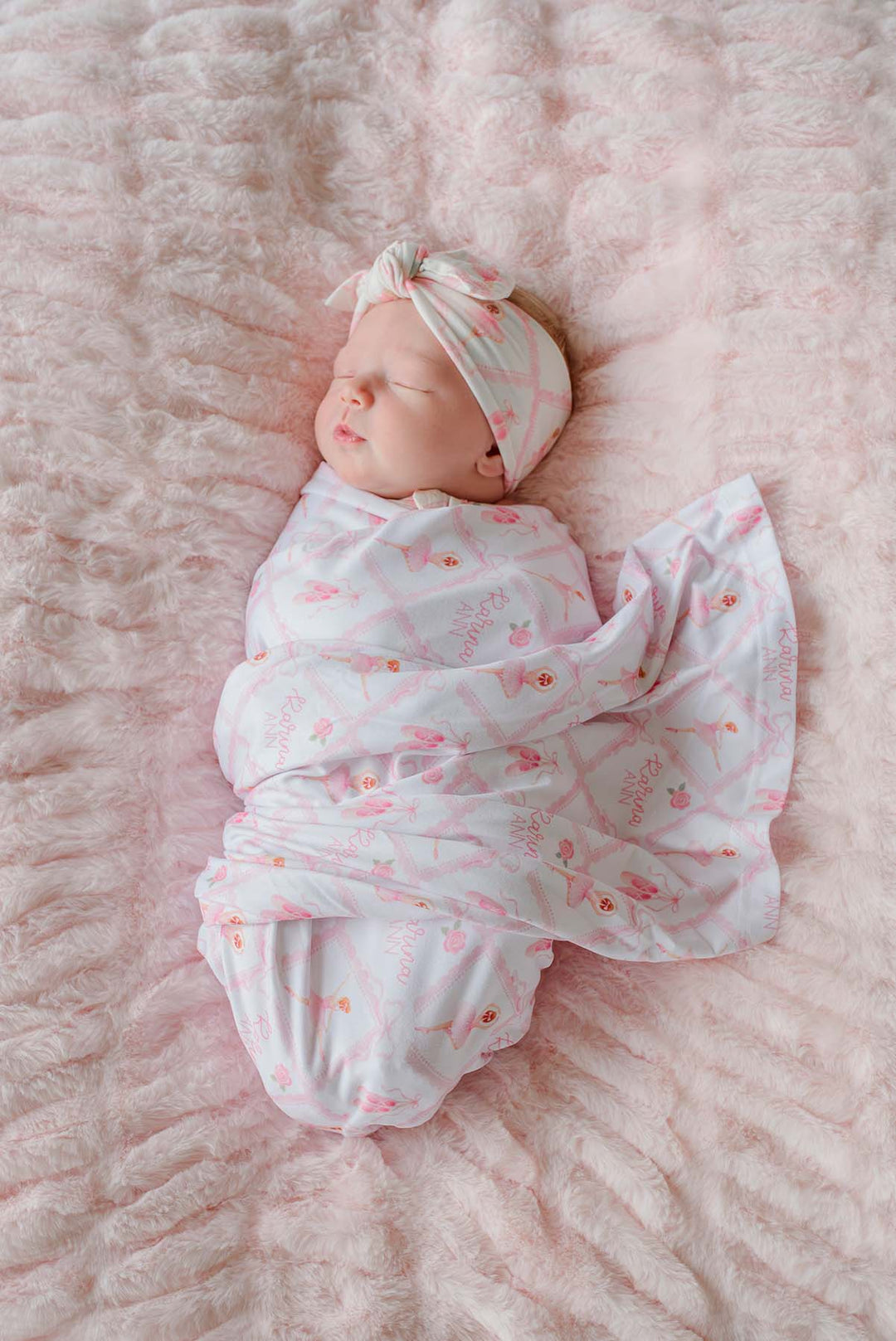 Sleeping newborn swaddled in a Caden Lane ballerina and roses pink print personalized swaddle blanket with matching knotted headband, on a pink fur blanket