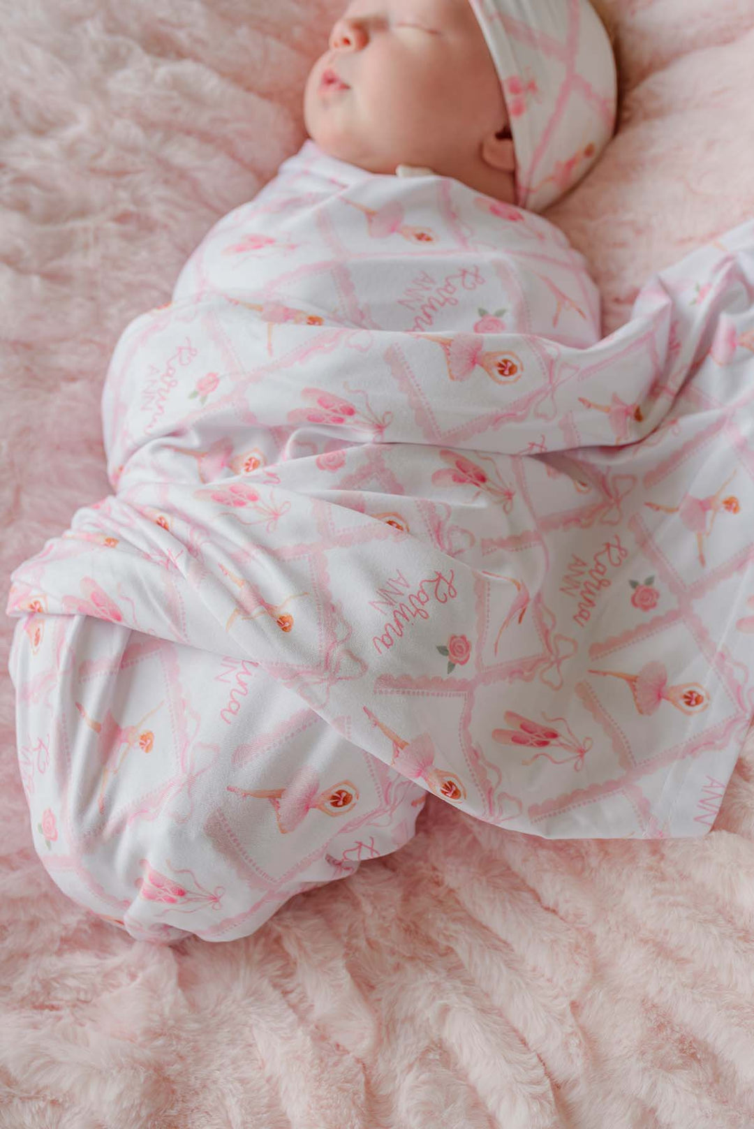 Sleeping newborn swaddled in a Caden Lane ballerina and roses pink print personalized swaddle blanket with matching knotted headband, on a pink fur blanket