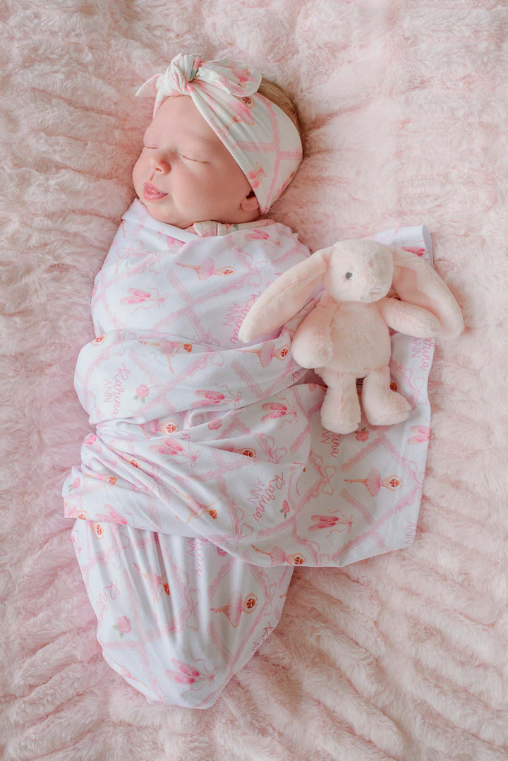 Sleeping newborn swaddled in a Caden Lane ballerina and roses pink print personalized swaddle blanket with matching knotted headband, on a pink fur blanket