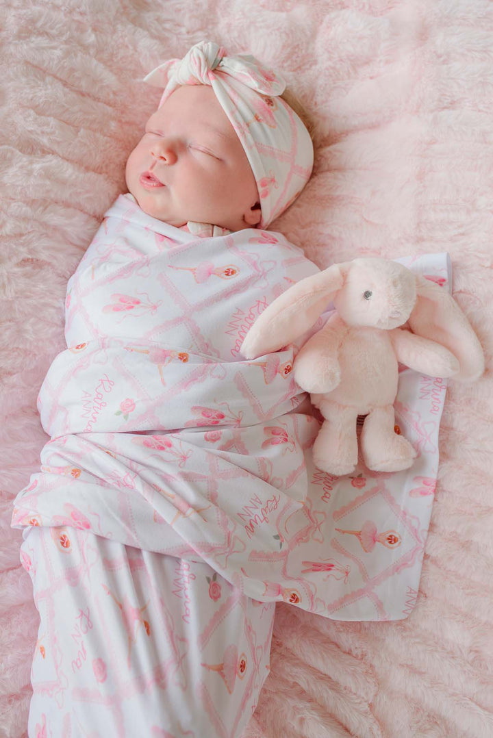 Sleeping newborn swaddled in a Caden Lane ballerina and roses pink print personalized swaddle blanket with matching knotted headband, on a pink fur blanket