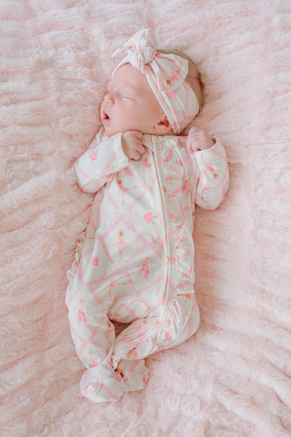 Sleeping newborn baby girl wearing a Caden Lane ballerina and roses pink print ruffle footie pajama with matching knotted headband, laying on a pink faux fur blanket