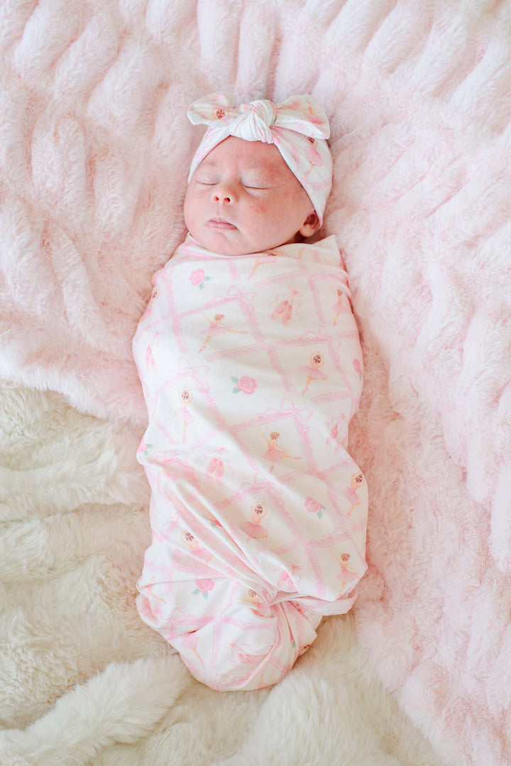 Close-up of sleeping newborn baby wearing a Caden Lane ballerina and roses pink print swaddle blanket with matching bow headwrap, laying on a pink faux fur blanket