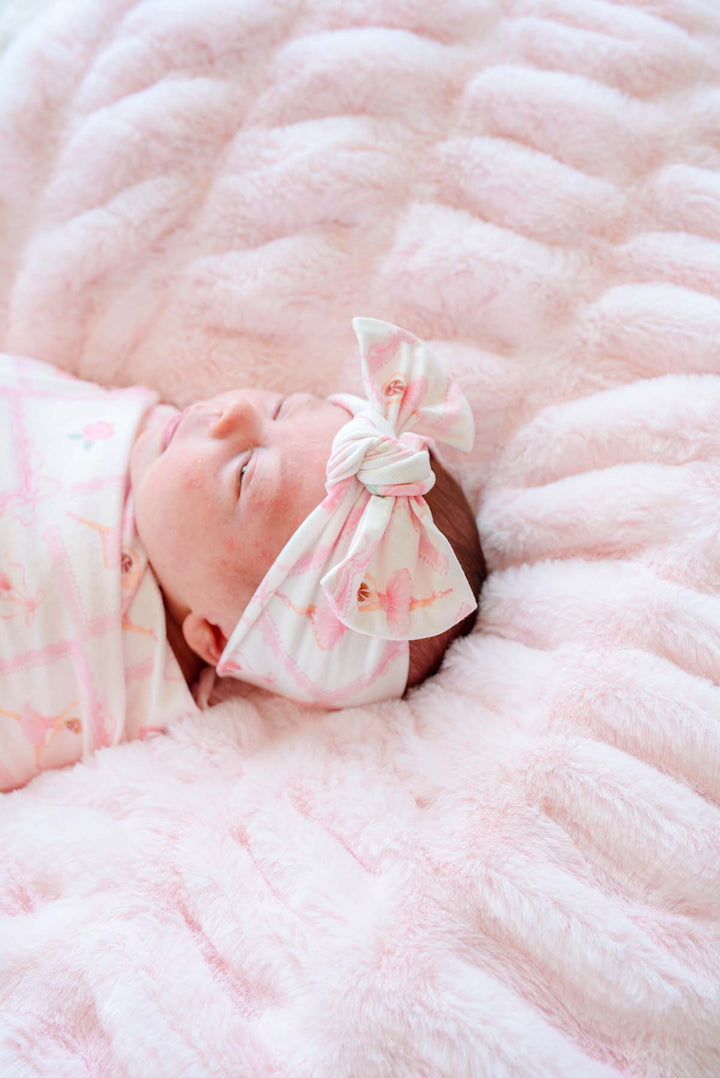 Close-up of sleeping newborn baby wearing a Caden Lane ballerina and roses pink print swaddle blanket with matching bow headwrap, laying on a pink faux fur blanket