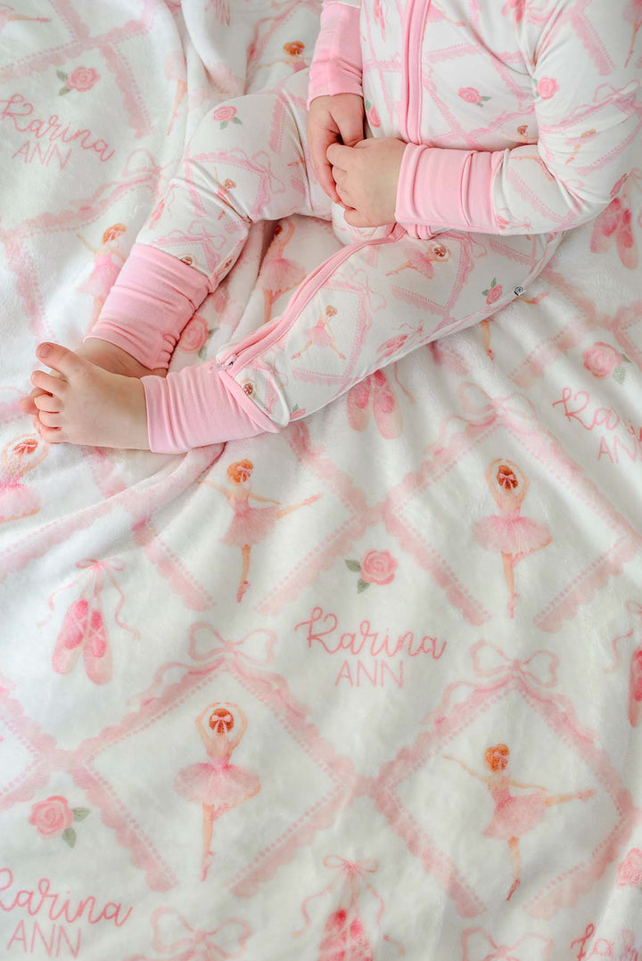 Baby in Caden Lane ballerina and roses pink print pajamas sitting with a matching personalized plush blanket embroidered with her name