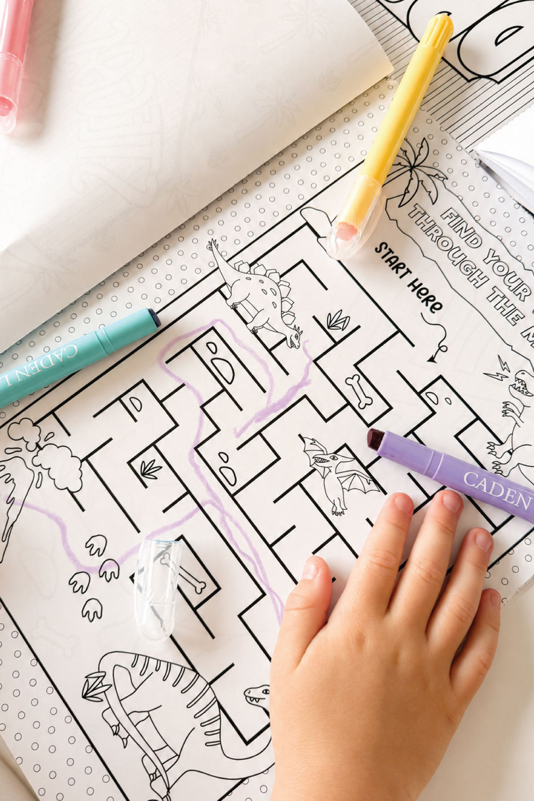 Child's hand coloring a dinosaur-themed maze page in the Caden Lane Rawr-Some Dinosaur coloring book with gel pens.
