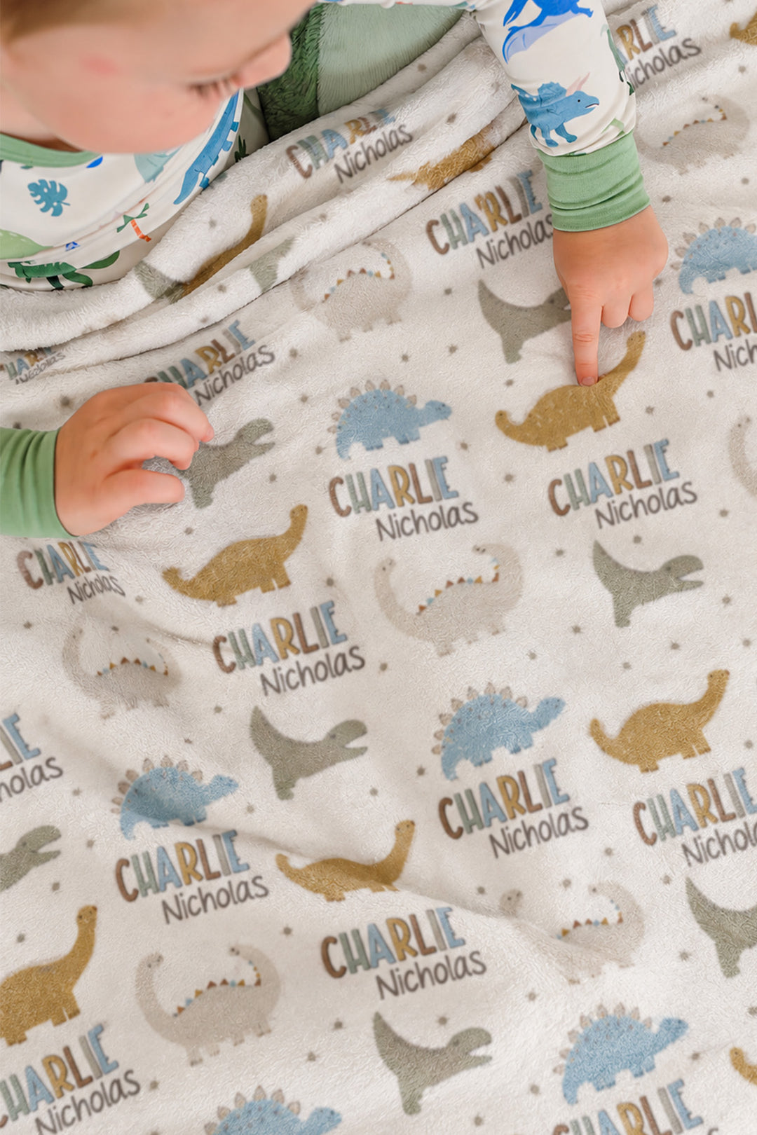 Close-up of two toddlers in dinosaur-print pajamas pointing at a personalized Caden Lane Dino Stomps blanket, showing dinosaur silhouettes and custom name print