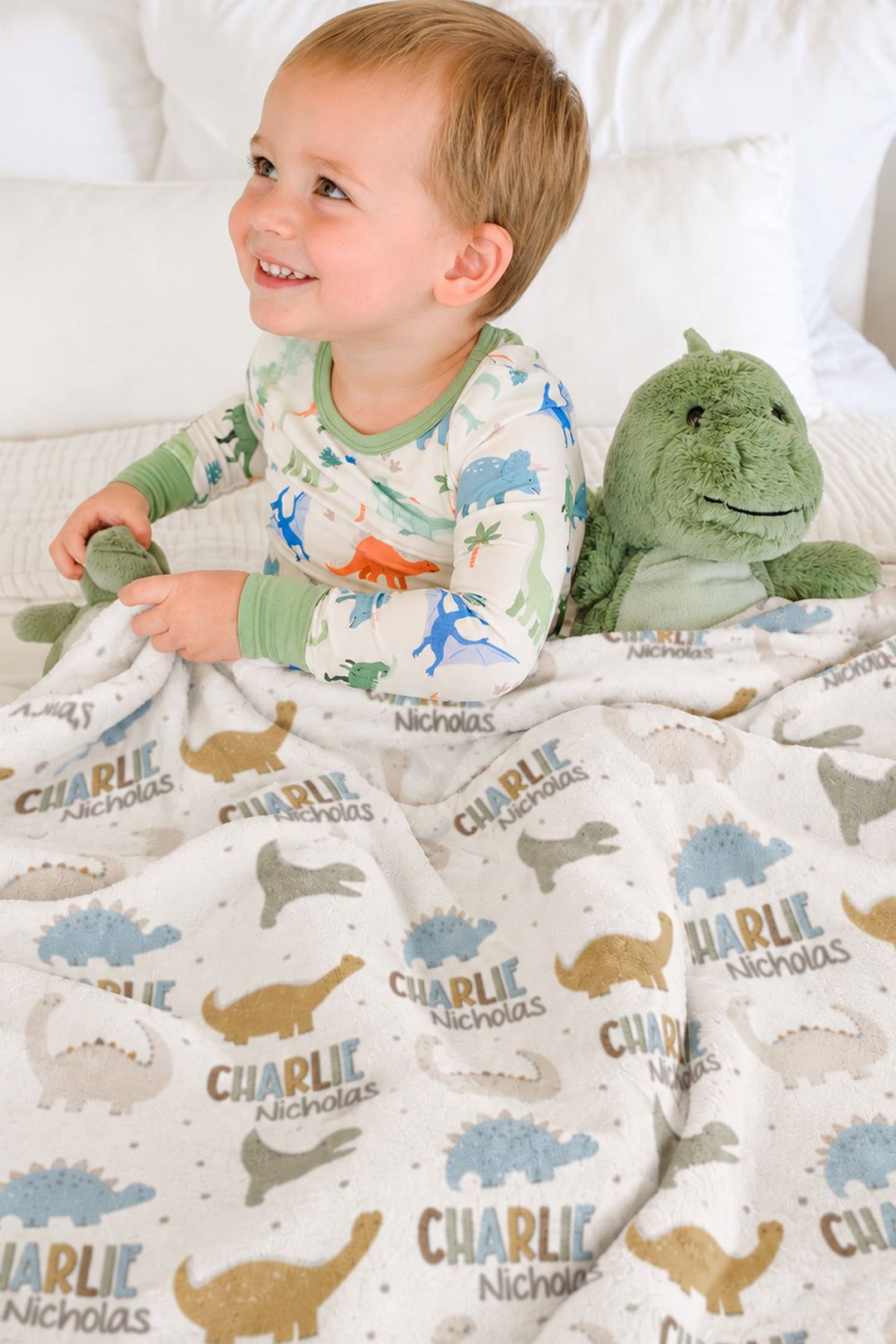 Smiling toddler in dinosaur-print pajamas sitting on a bed with a plush dinosaur toy, wrapped in a personalized Caden Lane Dino Stomps blanket