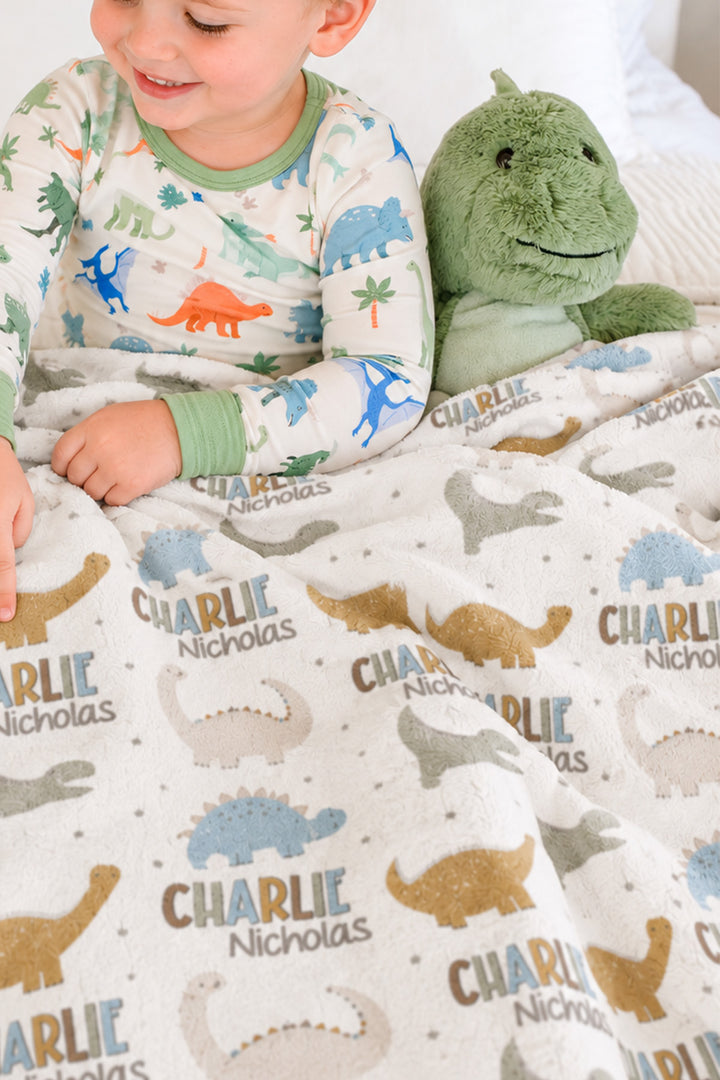 Toddler in dinosaur-print pajamas sitting on a bed next to a plush dinosaur toy, close-up view of a personalized Caden Lane Dino Stomps blanket with custom name print