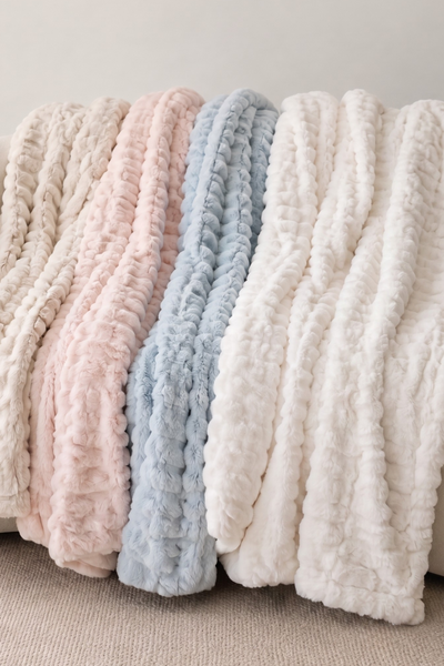 Three textured chunky knit throw blankets in cream, light pink, and light blue draped over a white curved armchair.
