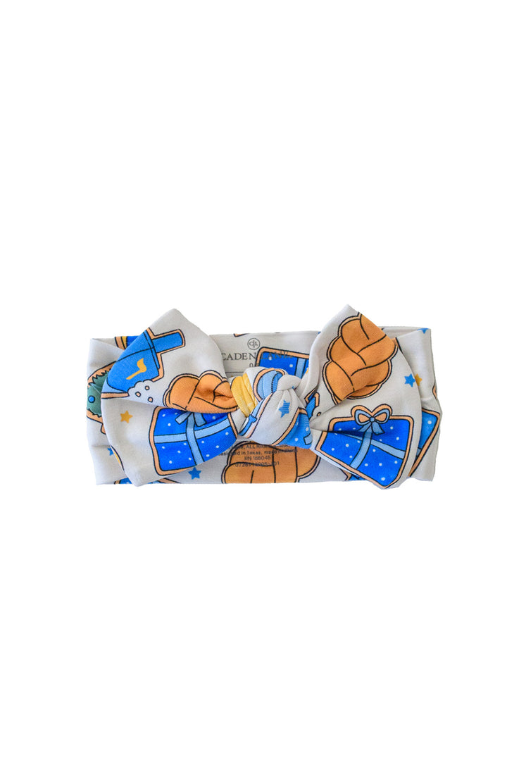Caden Lane Hanukkah baby headband with bow featuring blue gifts, dreidels, challah, and festive holiday print.