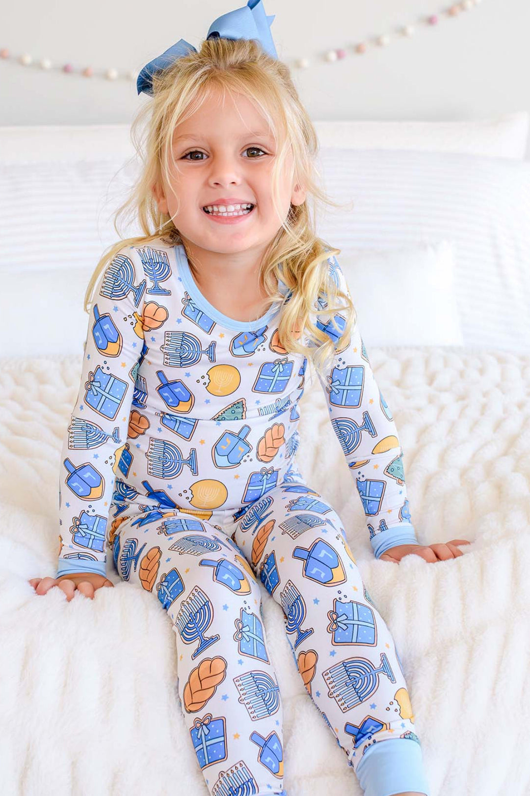 Young girl wearing Caden Lane Hanukkah pajamas with blue menorah, dreidel, gift, and challah print while sitting on a cozy white bed.