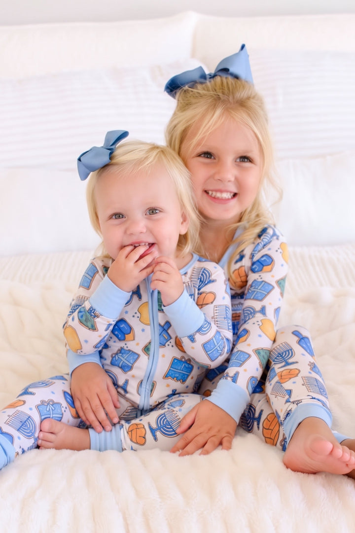 Two young children wearing coordinating Caden Lane Hanukkah pajamas with menorah, dreidel, gift, challah, and gelt designs.