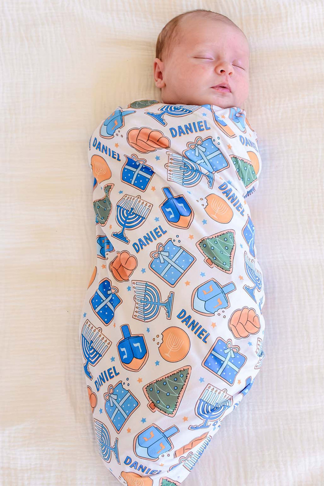 Sleeping newborn wrapped in a personalized Caden Lane Hanukkah swaddle featuring menorahs, dreidels, gifts, challah, gelt, and the name Daniel.