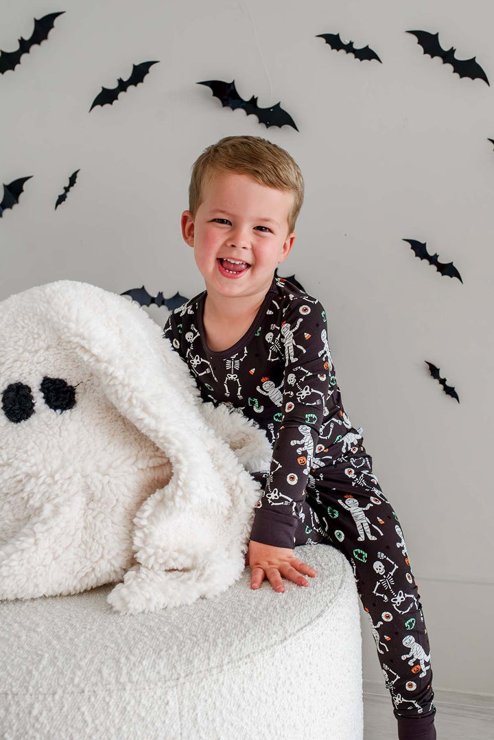 Boy leaning on plush ghost pillow and wearing a Caden Lane black two-piece pajama set with a white skeleton, mummy, and candy corn print, with Halloween bat decorations in the background