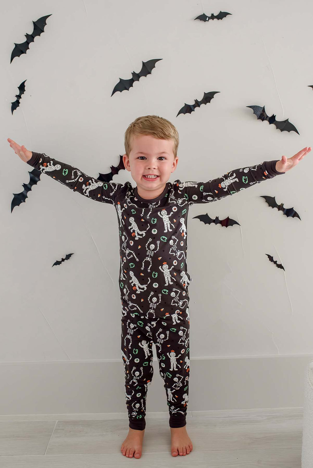Boy standing and wearing a Caden Lane black two-piece pajama set with a white skeleton, mummy, and candy corn print, with Halloween bat decorations in the background