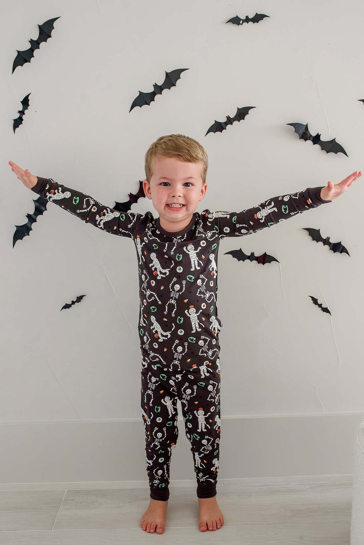 Boy standing and wearing a Caden Lane black two-piece pajama set with a white skeleton, mummy, and candy corn print, with Halloween bat decorations in the background