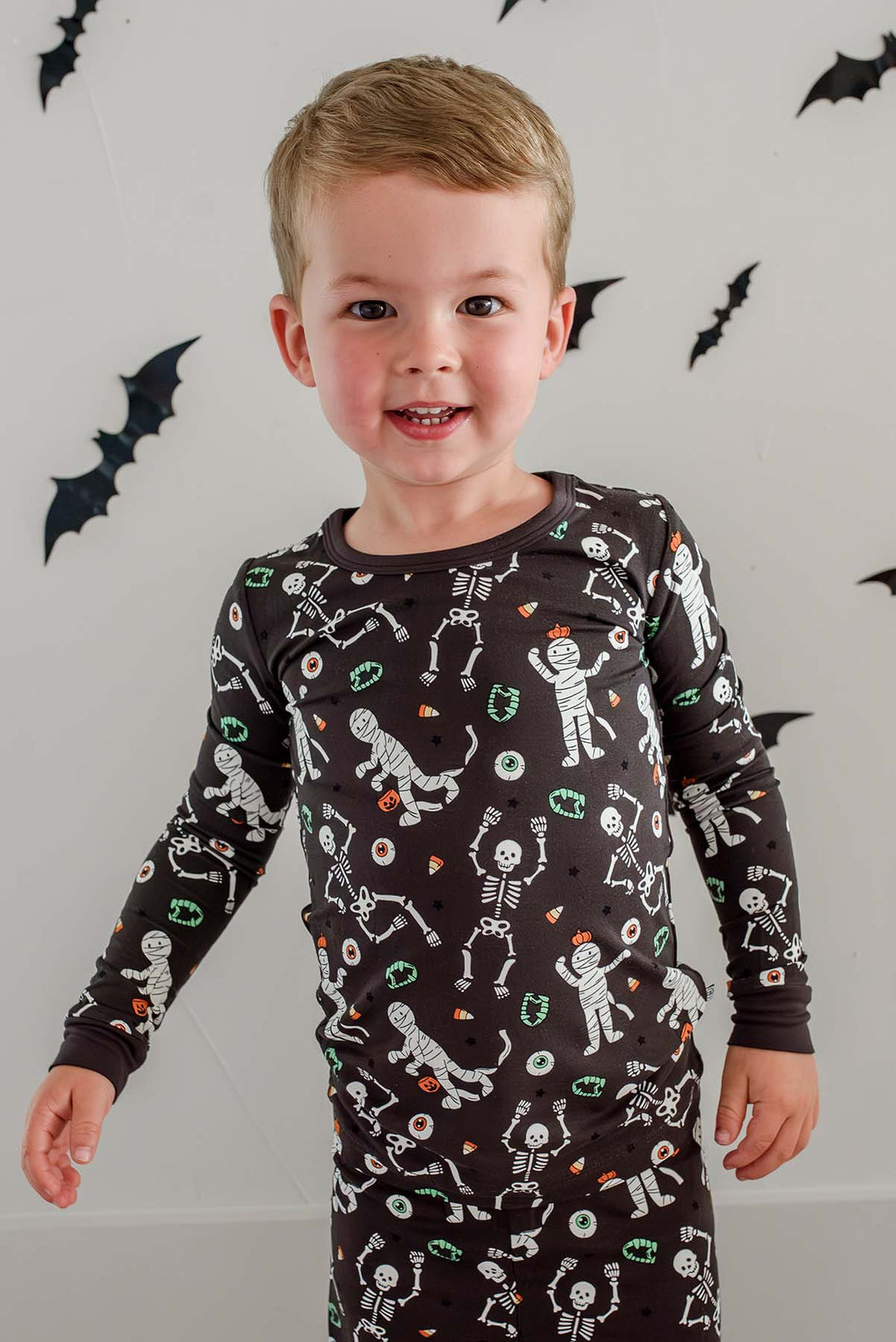 Boy standing and wearing a Caden Lane black two-piece pajama set with a white skeleton, mummy, and candy corn print, with Halloween bat decorations in the background
