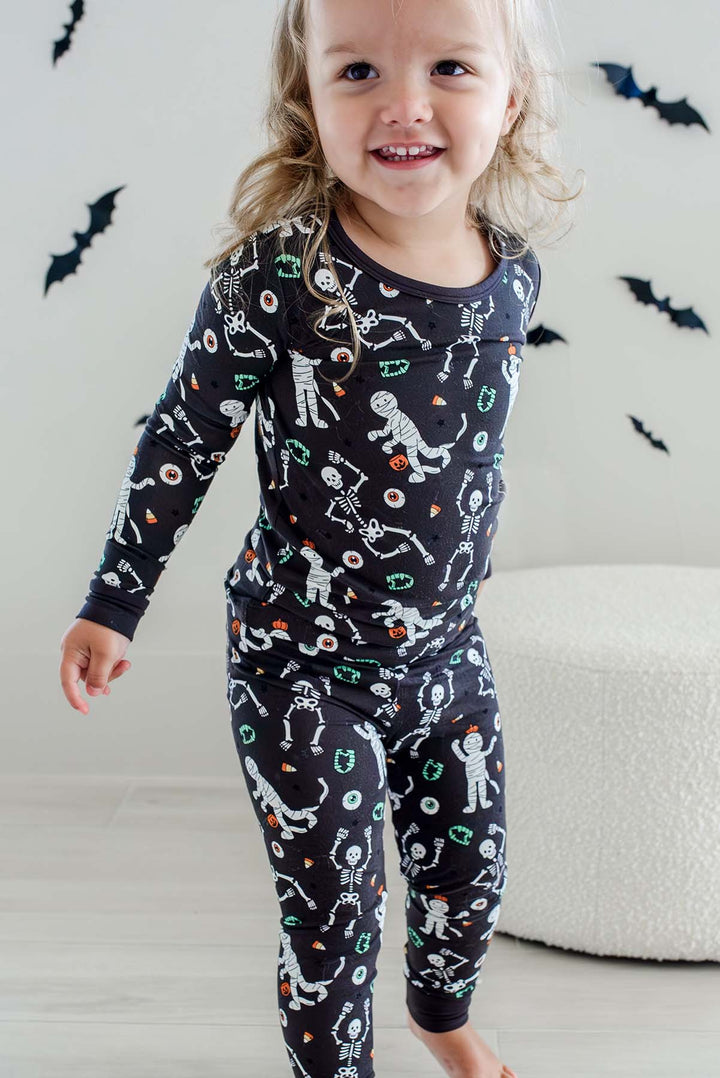 Girl standing and wearing a Caden Lane black two-piece pajama set with a white skeleton, mummy, and candy corn print, with Halloween bat decorations in the background
