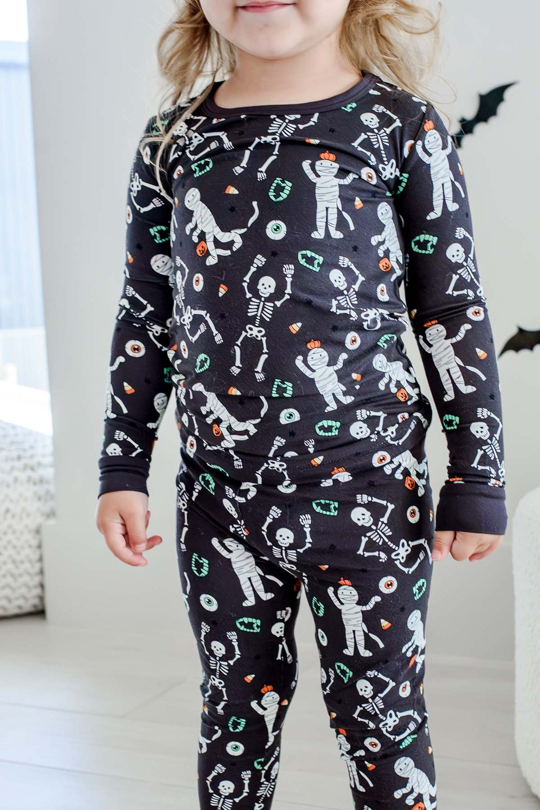 Young child standing and wearing a Caden Lane black two-piece pajama set with a white skeleton, mummy, and candy corn print, with Halloween bat decorations in the background