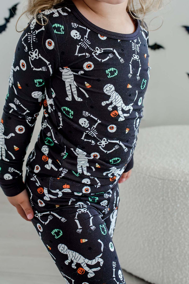 Young child standing and wearing a Caden Lane black two-piece pajama set with a white skeleton, mummy, and candy corn print, with Halloween bat decorations in the background