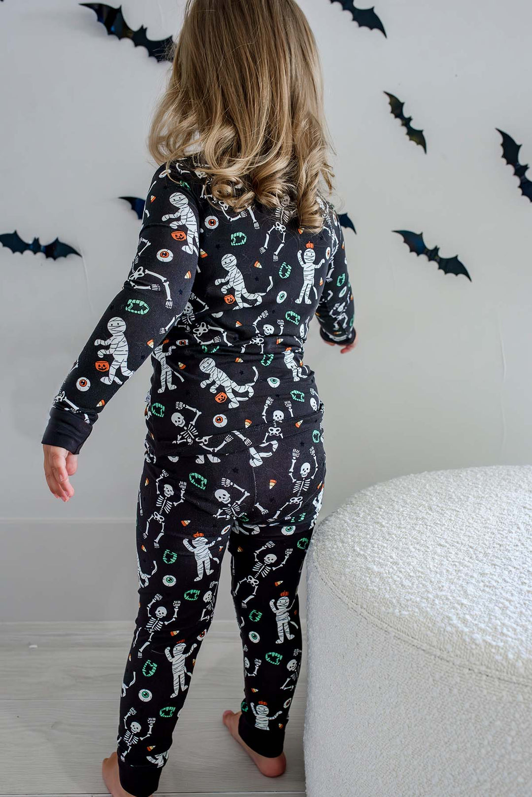 Girl standing and wearing a Caden Lane black two-piece pajama set with a white skeleton, mummy, and candy corn print, with Halloween bat decorations in the background