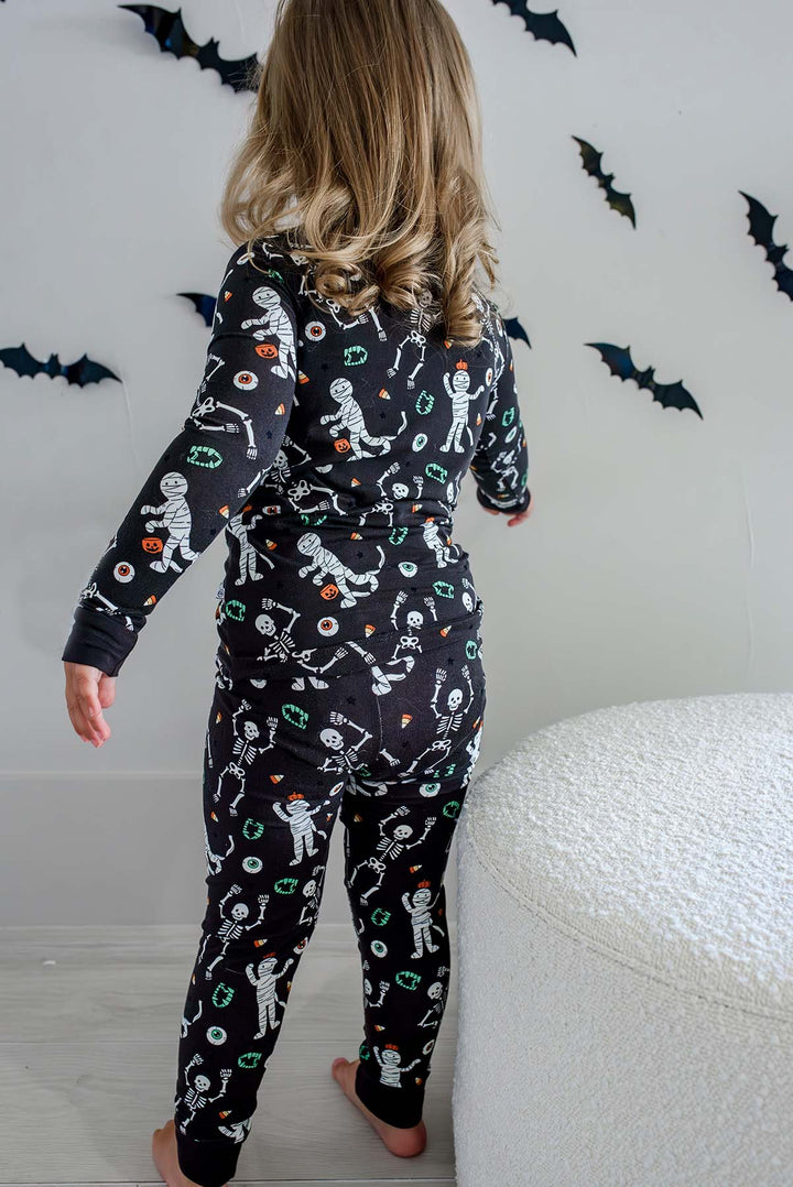Girl standing and wearing a Caden Lane black two-piece pajama set with a white skeleton, mummy, and candy corn print, with Halloween bat decorations in the background