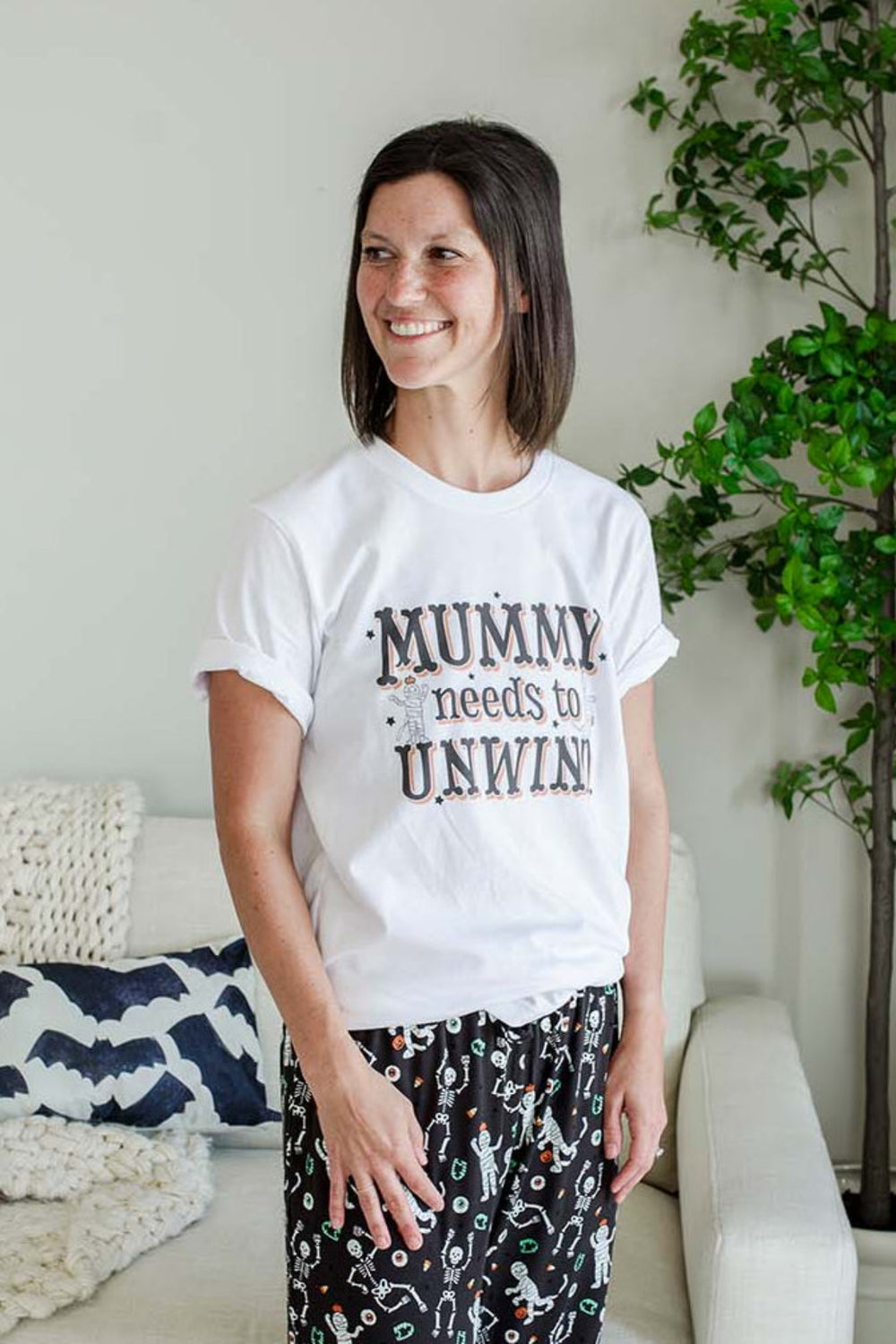 A woman stands in a living room wearing a white graphic T-shirt that reads 'MUMMY needs to UNWIND' and black patterned pajama pants. The room has a white sofa with a knit blanket, a potted tree, and a white plush ghost on the floor.